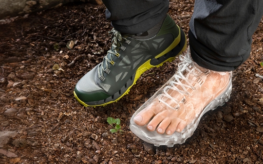 Auf einem Erdboden sind zwei Füße mit unterschiedlichem Schuhwerk zu sehen. Der rechte Fuß trägt einen Wanderschuh, während der linke Fuß denselben Schuh in einer komplett transparenten Version trägt, sodass der Fuß im Inneren sichtbar ist.